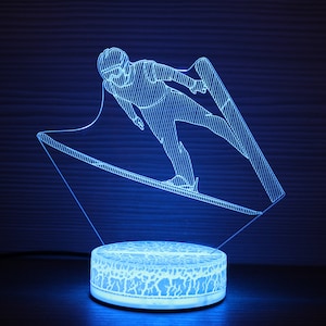 May include: A blue 3D printed acrylic night light depicting a skier in mid-air, jumping off a ski ramp. The light is on a white circular base with a textured surface.