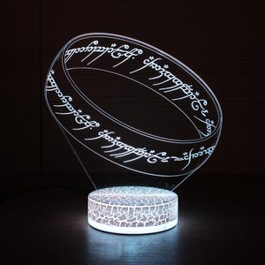 May include: A white acrylic lamp shaped like the One Ring from The Lord of the Rings. The ring has the inscription "One Ring to rule them all, One Ring to find them, One Ring to bring them all and in the darkness bind them" in Elvish script.