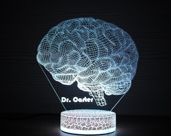 May include: A white 3D printed brain shaped night light with a textured base. The light is on and illuminates the brain and base. The text "Dr. Carter" is printed on the base.