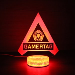 May include: A red triangular acrylic night light with a gamertag logo and a white base. The logo is a stylized design of a shield with a diamond in the center. The base has a decorative pattern.