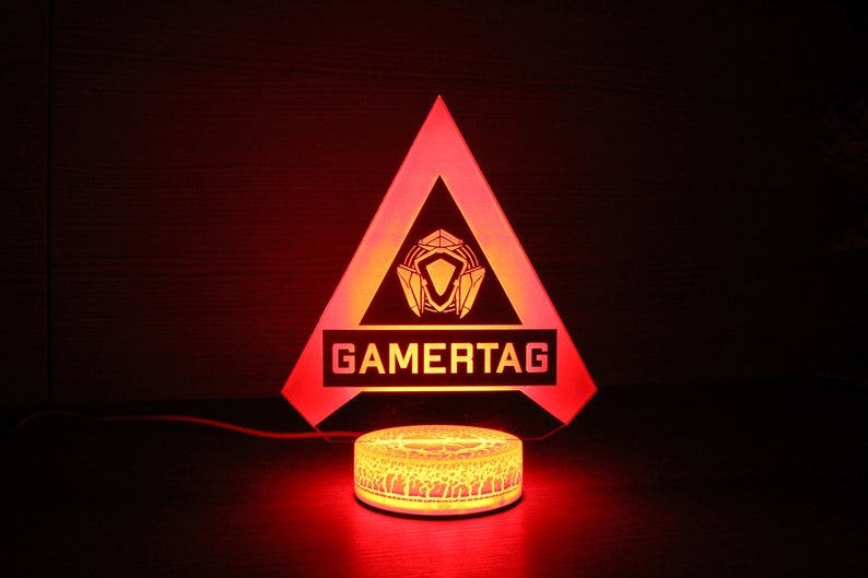 May include: A red triangular acrylic night light with a gamertag logo and a white base. The logo is a stylized design of a shield with a diamond in the center. The base has a decorative pattern.