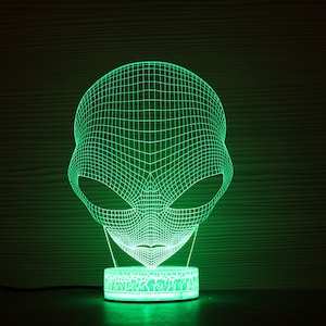 May include: A green, illuminated alien head lamp. The lamp has a 3D wireframe design of an alien head, emitting a bright green glow. The lamp sits on a round base with a textured pattern, and is plugged into a power source.
