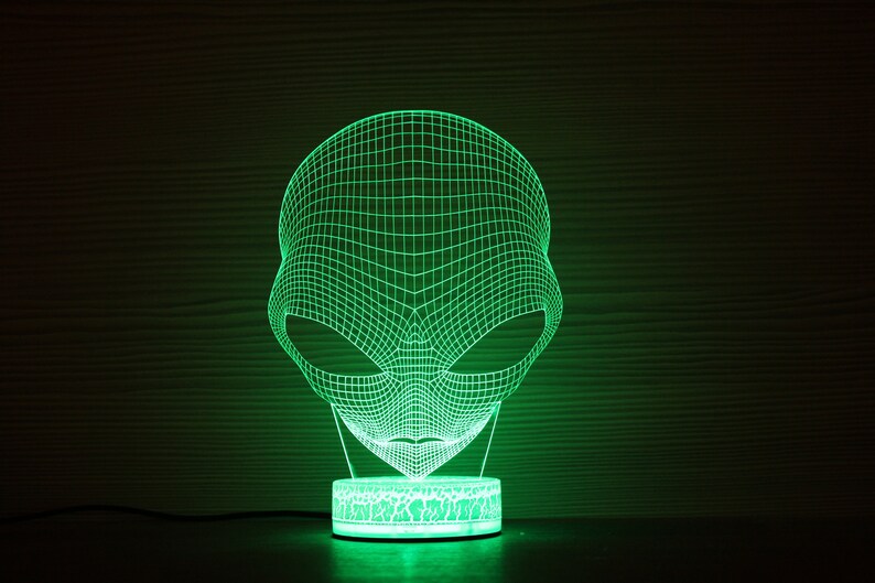 May include: A green, illuminated alien head lamp. The lamp has a 3D wireframe design of an alien head, emitting a bright green glow. The lamp sits on a round base with a textured pattern, and is plugged into a power source.
