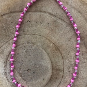 May include: A pink beaded necklace with the words "KISS YOU" spelled out in white letter beads. The necklace features a mix of light and dark pink beads, with clear beads interspersed throughout. The clasp is silver.