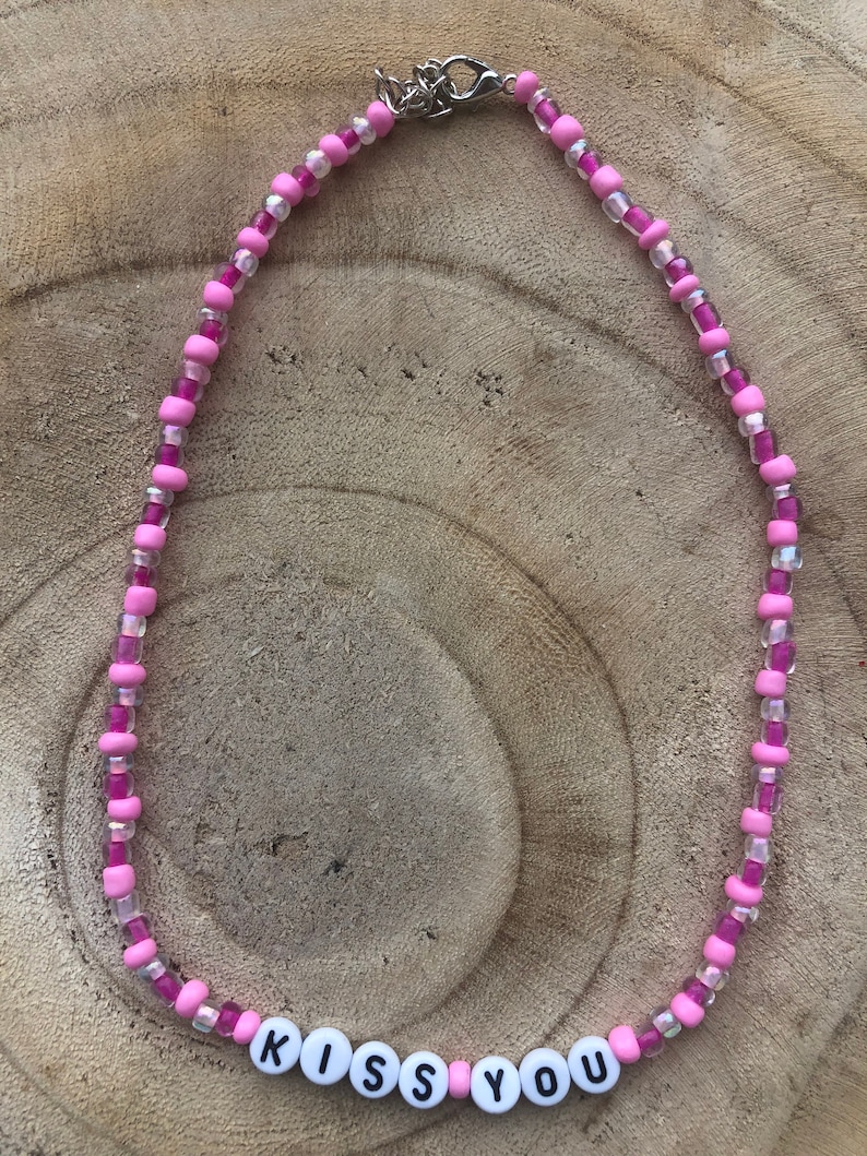 May include: A pink beaded necklace with the words "KISS YOU" spelled out in white letter beads. The necklace features a mix of light and dark pink beads, with clear beads interspersed throughout. The clasp is silver.