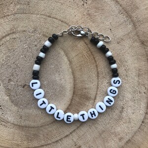 May include: A beaded bracelet with the words "LITTLE THINGS" spelled out in white letter beads. The bracelet also features black and white beads and a silver-colored clasp.