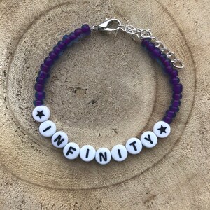 May include: A beaded bracelet with the word "INFINITY" spelled out in white letter beads. The bracelet features purple and blue beads and a silver-colored clasp. The bracelet is displayed on a wooden surface.