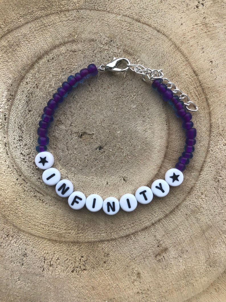 May include: A beaded bracelet with the word "INFINITY" spelled out in white letter beads. The bracelet features purple and blue beads and a silver-colored clasp. The bracelet is displayed on a wooden surface.