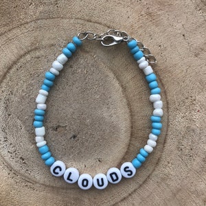 May include: A beaded bracelet with the word "CLOUDS" spelled out in white letter beads. The bracelet features alternating light blue and white beads, and a silver-colored clasp.