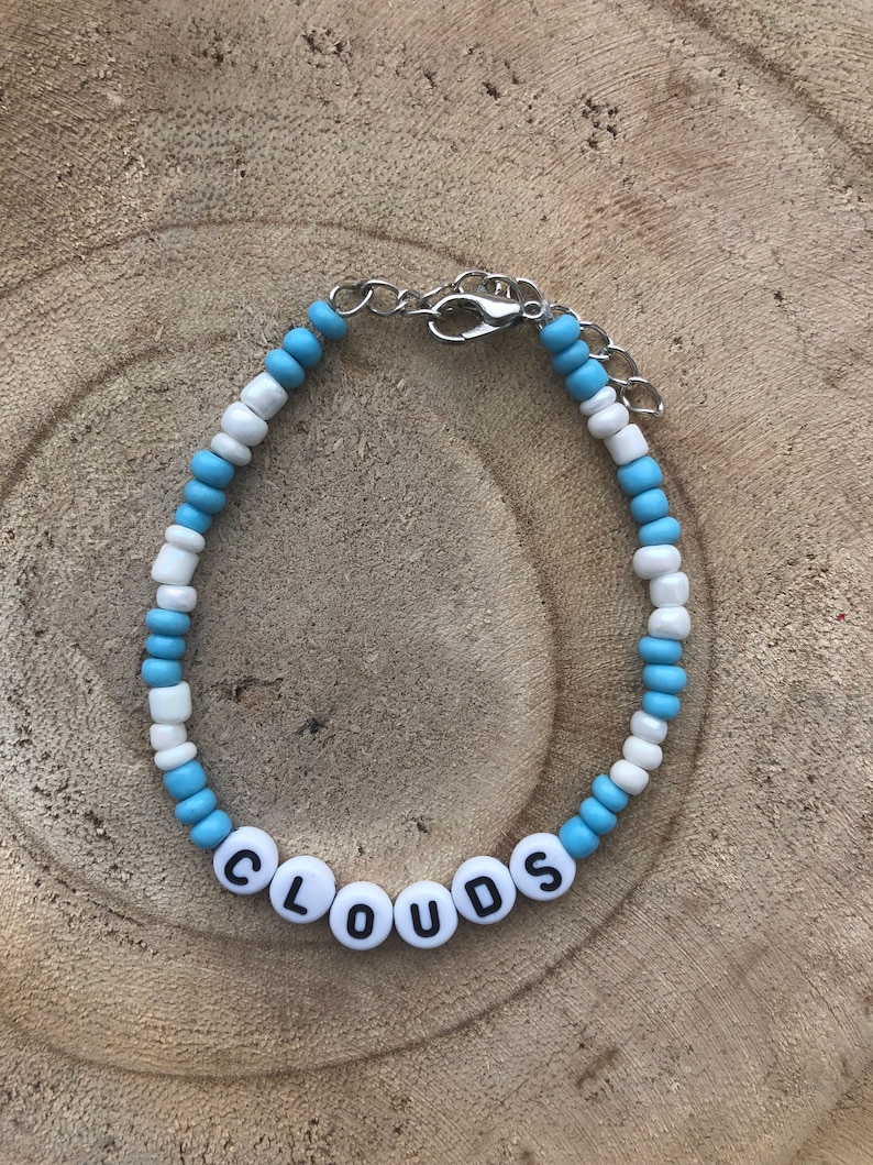 May include: A beaded bracelet with the word "CLOUDS" spelled out in white letter beads. The bracelet features alternating light blue and white beads, and a silver-colored clasp.