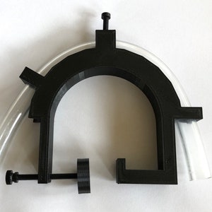 Puede incluir: Una abrazadera negra impresa en 3D con un diseño curvo, que sujeta un tubo de plástico transparente. La abrazadera tiene un tornillo para ajustar el agarre y está diseñada para asegurar el tubo. El fondo es blanco liso.