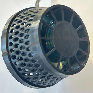 May include: A close-up of a black and silver cylindrical object with a perforated outer shell. The object has a circular end with a fan-like design. A curved metal hook extends from the top.