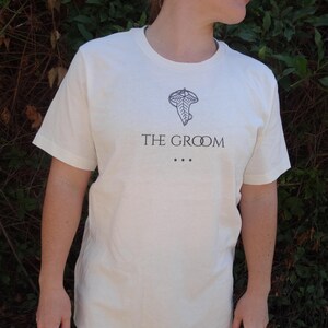 Puede incluir: Camiseta color crema con cuello redondo y mangas cortas. La camiseta presenta un gráfico negro de una hoja y el texto "THE GROOM" con tres puntos debajo. La camiseta está hecha de un material suave.