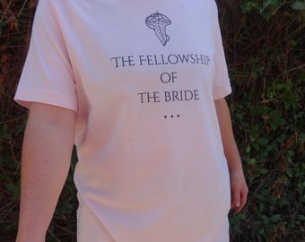 May include: A light pink t-shirt with the text "THE FELLOWSHIP OF THE BRIDE" in dark blue. Above the text is a stylized leaf design. The shirt is short-sleeved and appears to be made of cotton.