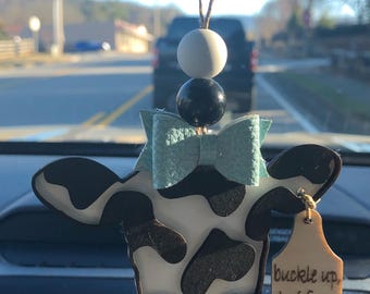 Puede incluir: Un ambientador de coche en forma de vaca blanco y negro con un lazo azul y una etiqueta de madera que dice "buckle up heifers".