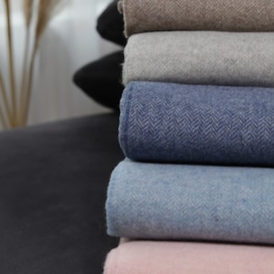 Può includere: Un mucchio di coperte piegate in vari colori, tra cui beige, grigio, blu e rosa. Le coperte sono fatte di un tessuto morbido e testurizzato.