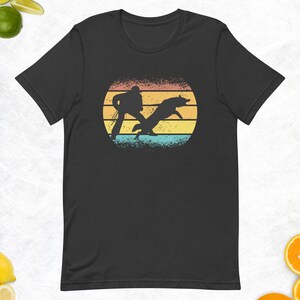 May include: A black t-shirt with a retro-style graphic of a person jumping over a dog in front of a sunset. The graphic is in shades of orange, yellow, and blue.