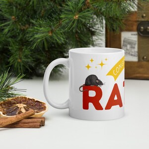 Peut inclure: Mug en céramique blanche avec une illustration de rat noir au-dessus des lettres rouges "RA". La tasse comporte également le mot "CONGRATS" en jaune et des étoiles dorées. Une idée cadeau amusante et unique.