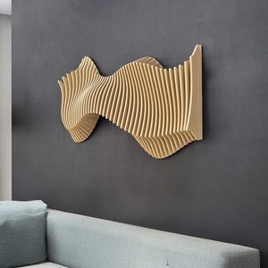 Puede incluir: Una obra de arte mural de madera con un diseño en forma de ola. La obra está hecha de listones de madera verticales y paralelos, creando un efecto texturizado y tridimensional. Está montada en una pared gris oscuro sobre un sofá azul claro.