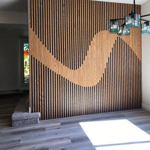 Könnte beinhalten: Eine Akzentwand aus Holz mit vertikalen schwarzen Streifen und einem wellenförmigen Design in natürlichen Holztönen. Die Wand ist Teil eines Raumes mit grauem Boden und einer dekorativen Leuchte.