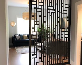 May include: A tall, rectangular room divider featuring a black metal frame with a geometric, open-work pattern of vertical and horizontal lines. The screen is positioned upright on a light-colored wooden floor, creating a semi-transparent partition.