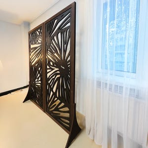 May include: A brown wooden room divider with a decorative cutout design. The divider is made of two panels, each with a different pattern. The panels are connected by a hinge.