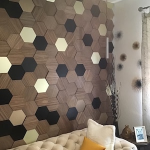 Può includere: Una parete rivestita con un motivo di piastrelle esagonali. Le piastrelle sono in legno e hanno una venatura del legno naturale. Alcune delle piastrelle sono dipinte di nero, mentre altre sono di colore oro chiaro. La parete è in un soggiorno e davanti c'è un divano.