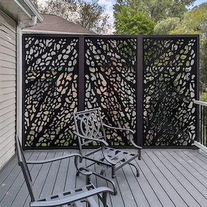 Pu&ograve; includere: Un paravento in metallo nero con un motivo organico ritagliato, installato su un terrazzo in legno. Due sedie in metallo sono di fronte allo schermo. Lo schermo &egrave; composto da tre pannelli, fornendo un elemento decorativo e funzionale.