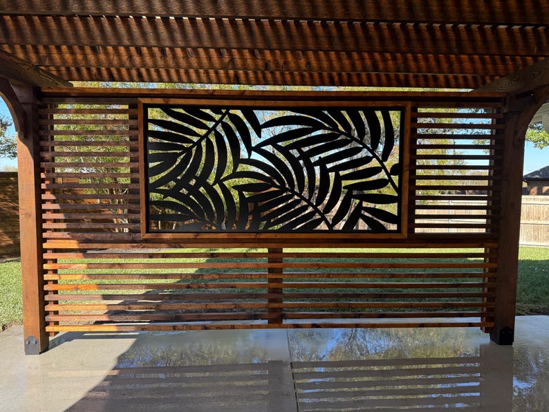 Outdoor pergola with a slatted wooden privacy screen featuring a tropical leaf cut-out design, casting shadows on the patio.