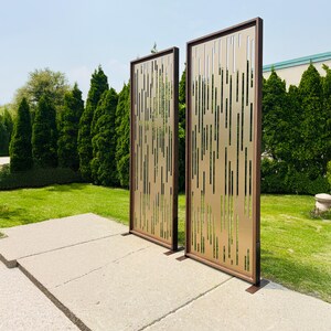 Pu&ograve; includere: Due schermi decorativi per esterni con finitura bronzo e design a taglio verticale. Gli schermi hanno una cornice marrone scuro e sono posizionati su una superficie di cemento, con erba verde e alberi sullo sfondo.