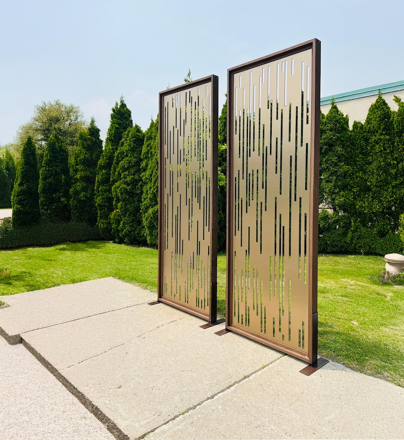 Pu&ograve; includere: Due schermi decorativi per esterni con finitura bronzo e design a taglio verticale. Gli schermi hanno una cornice marrone scuro e sono posizionati su una superficie di cemento, con erba verde e alberi sullo sfondo.