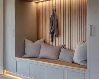 May include: A built-in bench with grey cabinets and a wooden seat. The back wall has vertical wooden slats and a hanging garment. Grey and pink cushions are arranged on the bench, with a warm light strip above and below.