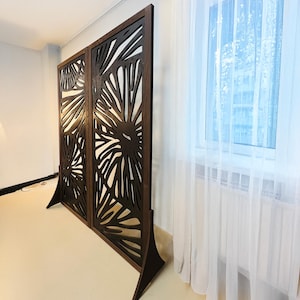 Brown wooden room divider with organic, abstract cut-out patterns, standing near a window with white sheer curtains