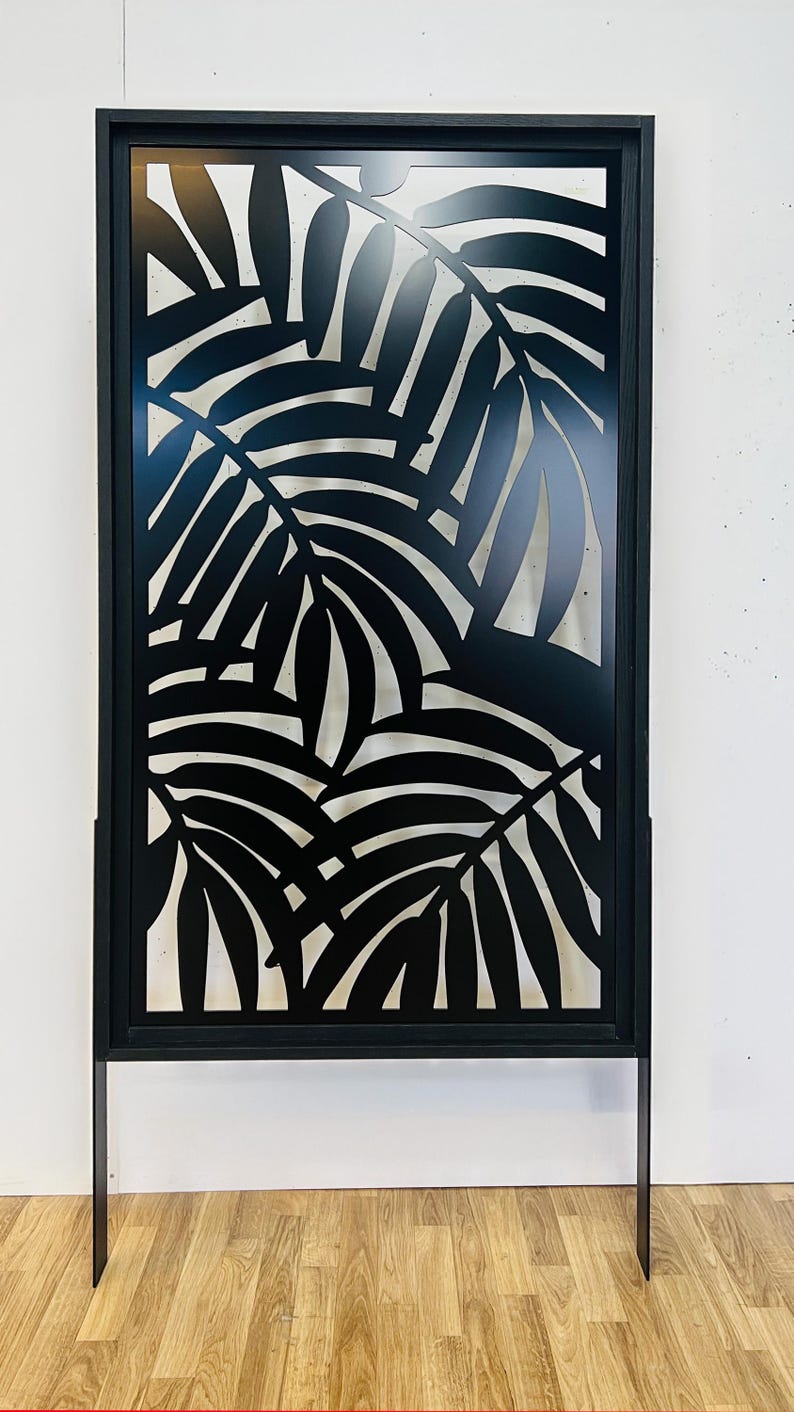 Pu&ograve; includere: Schermo decorativo in metallo nero con un design a foglie di palma ritagliate. Lo schermo rettangolare ha una cornice nera e poggia su due gambe in metallo nero. Il design crea un contrasto tra le foglie nere e lo sfondo.