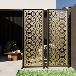 Pu&ograve; includere: Due schermi decorativi per esterni con un motivo geometrico. Gli schermi hanno una cornice marrone scuro e un design traforato color oro. Gli schermi sono su un patio di cemento con un pezzo di erba verde in primo piano.