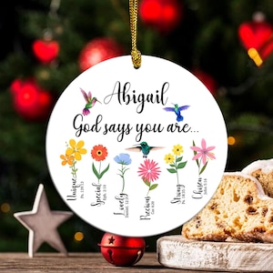 May include: A white round ornament with a gold string for hanging. It has a hummingbird and flowers with bible verses around the edge. The text reads "Abigail God says you are... Unique, Special, Lovely, Precious, Strong, Chosen".