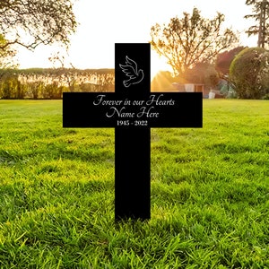K&ouml;nnte beinhalten: Schwarzes Holzkreuz mit einer wei&szlig;en Taube und dem Text "Forever in our Hearts Name Here 1945 - 2022" auf einem gr&uuml;nen Rasen.