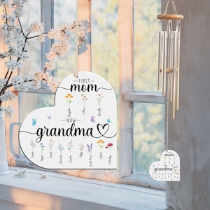 May include: White heart-shaped wooden sign with the text "First Mom - Now Grandma" and a floral design. The sign is hanging on a window with a silver wind chime hanging next to it.