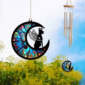 May include: A stained glass moon wind chime with a black silhouette of a dog with angel wings. The dog is sitting on the moon and has a heart with the name "Leo" written below it. The wind chime is hanging from a white string.