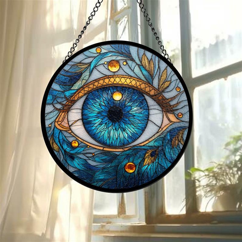 May include: Round stained glass window art featuring a detailed blue eye design. The eye is surrounded by blue and gold leaf patterns, with a black metal frame and hanging chain. The window art is displayed in front of a window.