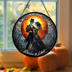 May include: Round stained glass window art featuring a skeleton couple in formal attire, embracing in front of a vibrant orange sunset. The artwork includes the names "James & Lilah" and is suspended by a chain. Pumpkins are visible in the background.