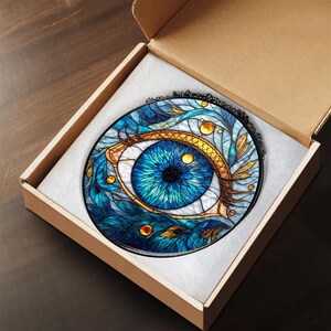 May include: A round, stained-glass style pendant featuring a detailed blue eye design with gold and teal accents. The pendant is set in a black frame and comes in a cardboard box with a white interior. The eye design is intricate and artistic.