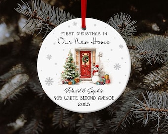May include: A white Christmas ornament with a red ribbon. The ornament features a festive scene with a red door, Christmas tree, snowman, and gifts. Text reads "First Christmas in Our New Home," names, and address.