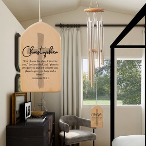 May include: A wooden wind chime with a personalized inscription that reads "Christopher" and a Bible verse from Jeremiah 29:11. The wind chime is hanging from a ceiling and has a cross design.