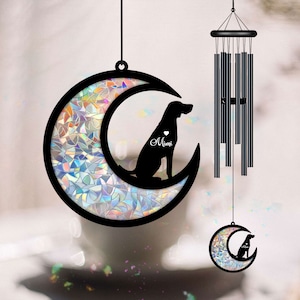May include: Black metal dog silhouette wind chimes. The dog is sitting inside a crescent moon with iridescent glass. The name "Mimi" is written on the dog's chest. The wind chimes have a black top and multiple black chimes.