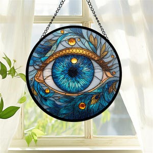 May include: Round stained glass art piece featuring a detailed blue eye design. The eye is surrounded by blue and gold accents, with a black border and a hanging chain. The artwork is displayed in front of a window.