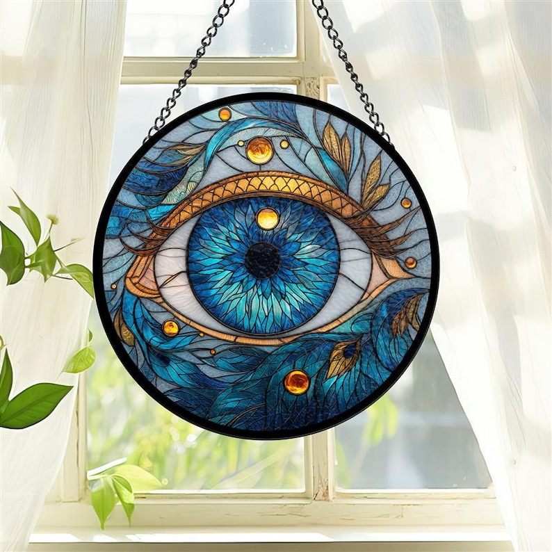 May include: Round stained glass art piece featuring a detailed blue eye design. The eye is surrounded by blue and gold accents, with a black border and a hanging chain. The artwork is displayed in front of a window.