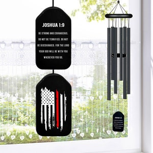 May include: Black wind chime with a red, white, and blue American flag design and a black tag with the text "Joshua 1:9" and a Bible verse. The tag is hanging from the top of the wind chime.