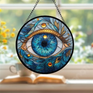 May include: Round stained glass art piece featuring a detailed blue eye design. The eye is surrounded by blue and gold floral elements, with a black border and hanging chain. The artwork is displayed in front of a window.