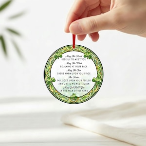 May include: A round, decorative ornament with a green Celtic knot design and shamrocks. The ornament has a white center with the text "May The Road Rise Up To Meet You" and other Irish blessings. It is suspended by a red ribbon.